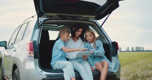 Woman and Her Teen Son and Daughter Sitting in Auto Trunk, Stopped in the Field During a trip