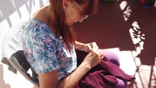 Older Caucasian woman sews her home clothes on a balcony with sunshine, portrait view