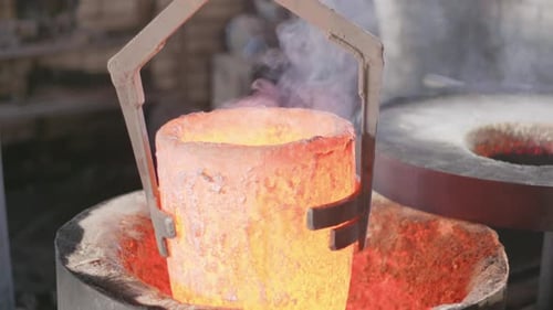 Slow motion hoist of red hot melting pot from furnace in small foundry industry setting