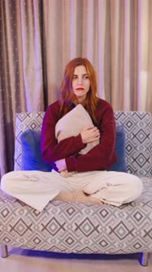 A Sad Young Redhaired Woman Sits Alone on a Couch Hugging a Pillow Tightly Lost in Her Thoughts A