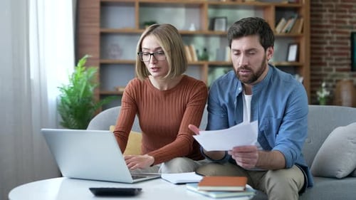 Unhappy shocked married couple looking at large utility bills sitting at home office.
