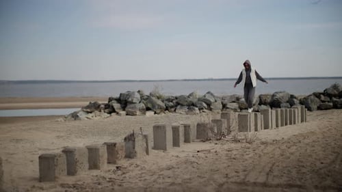 Woman in a Hooded Jacket Walking Across Concrete Blocks By a Sandy Beach on a Cloudy Day