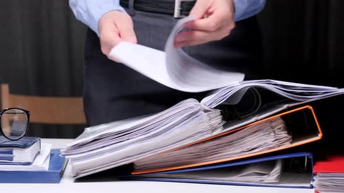 Office Worker is Searching Through a Large Stack of Binders and Documents