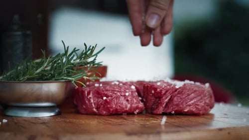 BBQ in the garden. Woman seasons filet mignon resting on the table with pepper and rosemary.