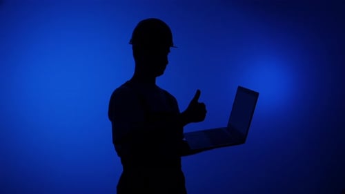 Silhouette of Worker in Hard Hat Holding a Laptop and Giving a Thumbs Up Against Vibrant Blue
