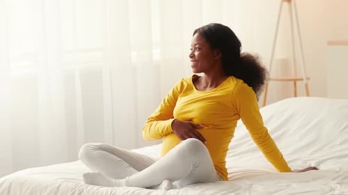 Pregnant woman sits in sunny bedroom and smiles