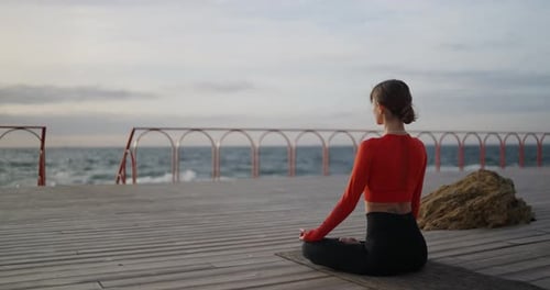 Rear View of a Happy Brunette Girl Practicing Yoga with an Orange Sweater Sitting on the Carpet and