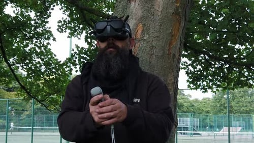 Bearded male wearing FPV virtual reality goggles controlling drone in park with motion joystick