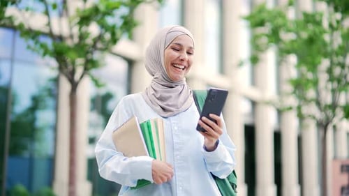 Smiling muslim female student in hijab with books in hands using smartphone standing in campus space