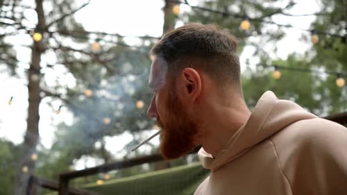 Un jeune homme barbu aux cheveux roux allume une cigarette sur fond d'arbres Le gars fume