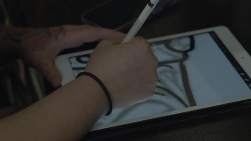 close up shot of a woman's tattooed hands as she uses a stylus to draw a design on an electronic tab
