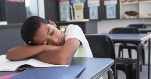 In school, in the classroom, a young biracial student is yawning at his desk with copy space