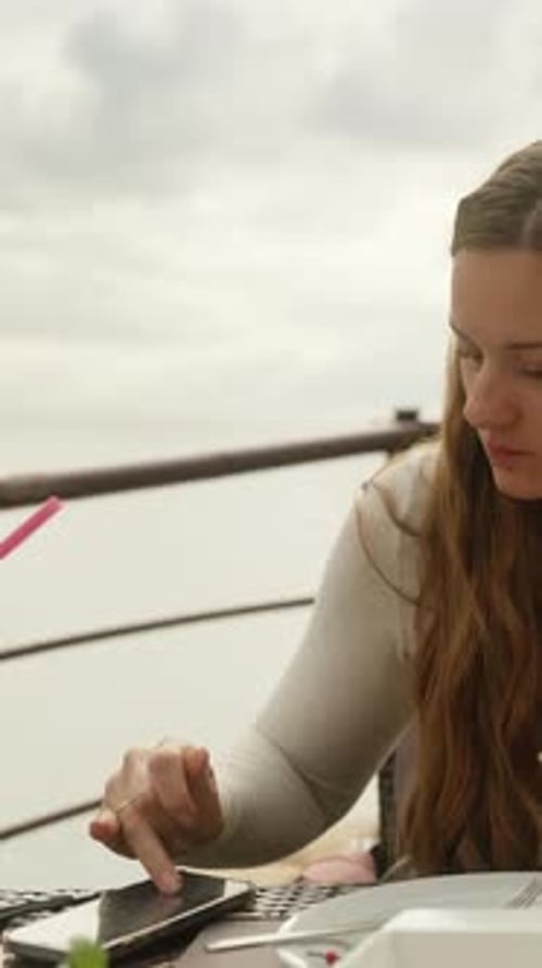A young woman eats pizza in a restaurant on a cliff by the sea and writes something on her phone. Ve