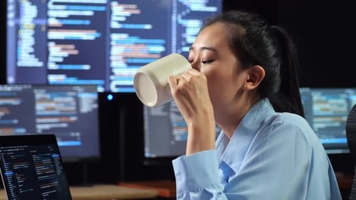 Une programmeuse asiatique boit du café tout en écrivant du code sur un ordinateur portable à l'aide de plusieurs moniteurs