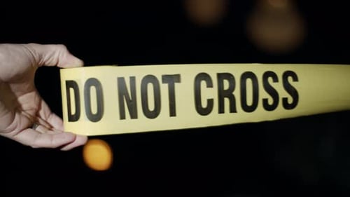 Close-up of a police officer's hand as he rolls out crime scene tape. Flashing emergency lights can