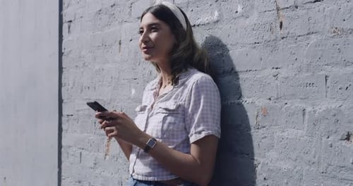 Happy woman sending a text message on her phone in the city against a grey wall