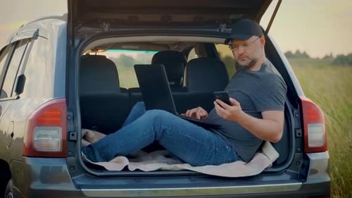 Homem de negócios adulto usando seu laptop e smartphone sentado no porta-malas do carro enquanto viaja trabalhando