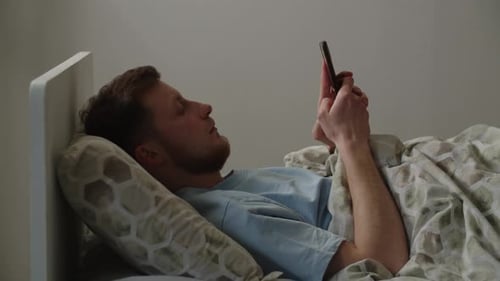 A Young Man Lies on a Bed and Browses Social Networks on a Smartphone A Man in a Blue Tshirt Looks