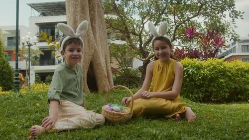 Happy Children Wearing Bunny Ears Sitting on Grass with Basket of Colorful Easter Eggs in Garden