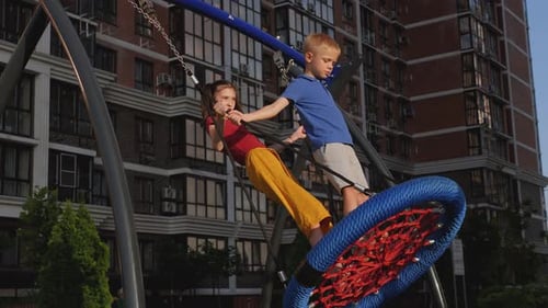 Little Boy and Girl Ride Together on a Swing in a Modern Yard and Playground