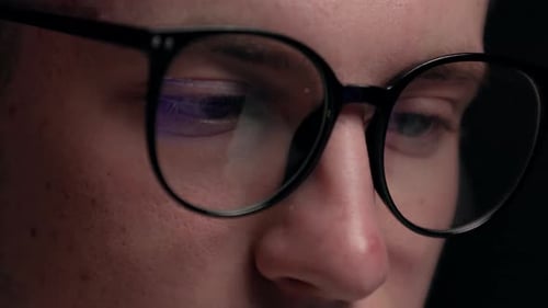 Close up focused face of programmer in computer glasses with monitors reflection works online late.