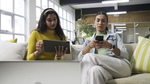 Diverse women colleagues comparing tablet charts, printed graphs at office lounge after phone alert