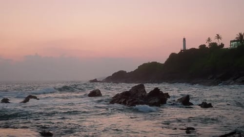 Sunset Paints Sky in Soft Pink Hues By a Hillside Lighthouse Waves Crash Against Rocky Shore