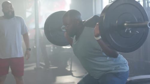 African American man doing barbell back squats bracing and training at gym with spotter watching