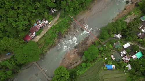 A wide aerial angle settles over a forested valley with a rocky river and red bridge centered in