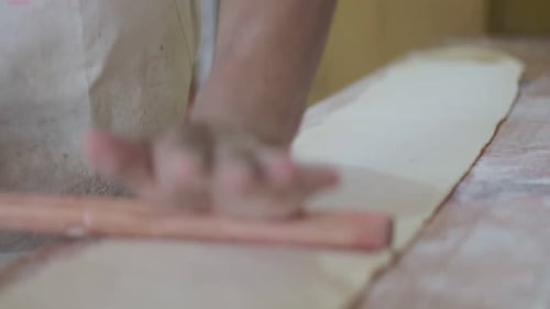 Close-up of a pastry chef rolling dough with hands using a wooden rolling pin on a work surface.