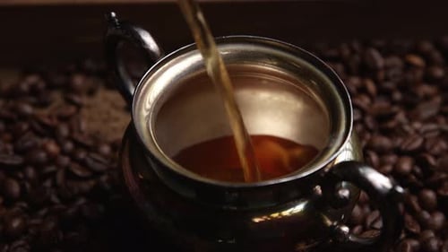 Luxurious rich coffee pouring into ornate silver cup on dark ground aromatic coffee beans close up