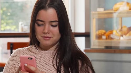 Cheerful Young Woman Smiling While Using Her Smartphone at a Cozy Coffee Shop
