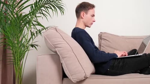 A Man Lies at Home on a Beige Sofa and Works with a Laptop in His Hands