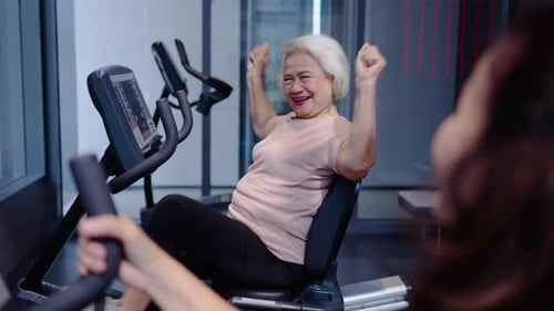 Happy senior Asian woman exercising on recumbent bike at gym, smiling and cheering with trainer.