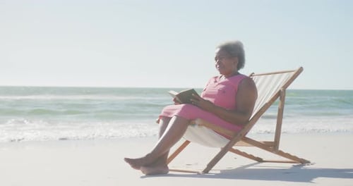 Happy senior african american woman sitting on deck chair and reading book at beach, in slow motion