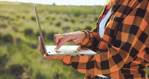 Close up of farmers woman hands taking notes and searches for information in laptop in agricultural