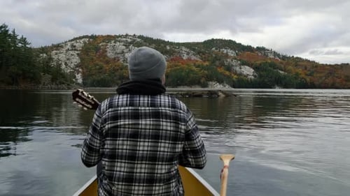 Man plays Guitar in Canoe in Autumn Leaf Color Forest on Canada lake. Wide Pan of Fall Trees, Rock C