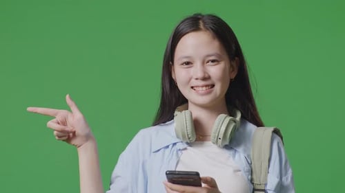 Asian Teen Girl Student With A Backpack Using A Smartphone And Pointing To Side In The Green Screen
