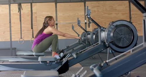 Determined unaltered biracial woman exercising on rowing machine at gym, in slow motion