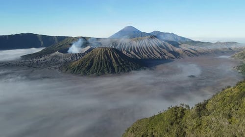 Aerial drone footage of Mount Bromo volcano with smoke rising, surrounded by misty sea of sand and