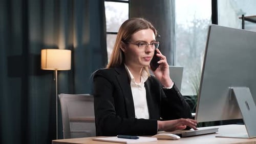Smiling Young Business Woman Talking on Phone Using Laptop Sits at Office Desk