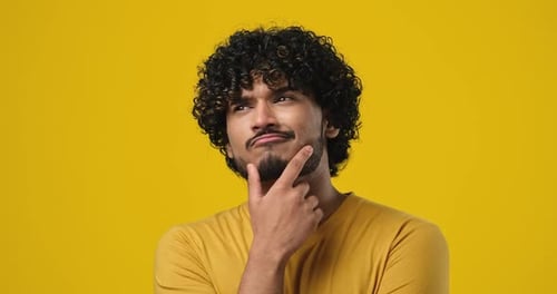 Pensive Young Indian Man Thinking with Hand Near Chin Standing Over Orange Studio Background