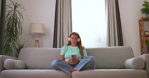 Frau in weißen Kopfhörern mit Smartphone hört zu Hause Musik auf dem Sofa