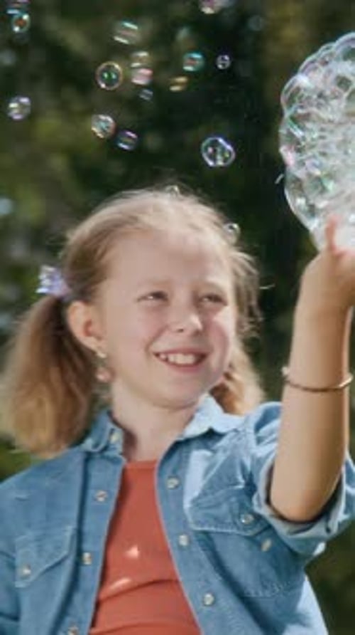 Vertical of Happy Gen Alpha Girl Catching Soap Bubbles in Sunny Weather Outdoors