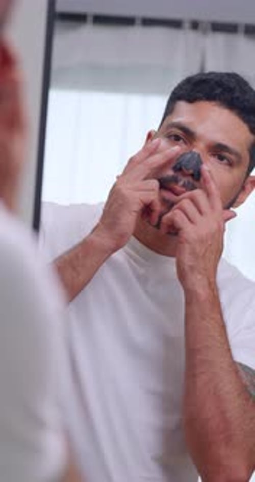 Handsome Asian man uses a nose patch to remove blackheads while standing in front of a mirror.