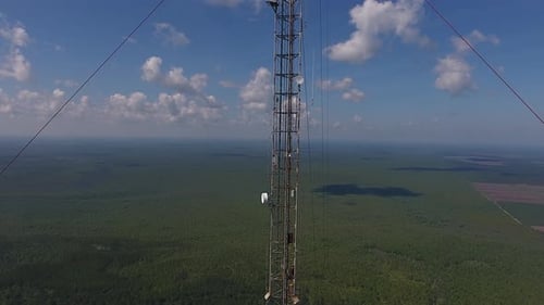 Drone shot moving away from a communication tower. A forest surrounds the tower below. The blue sky
