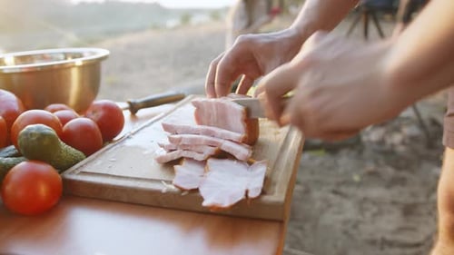 A Man Cuts Bacon on a Table with Tomatoes and Cucumbers for a Picnic with Friends