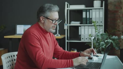 Mature Engineer Working on Wind Energy Project on Laptop at Office Desk