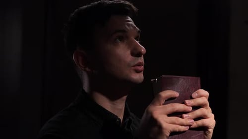 Religious Man Praying and Holding Holy Bible in Dark Room