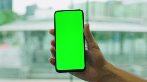 Closeup of a Male Hand Holding a Smartphone with a Green Screen on a Blurred City Background Mock Up